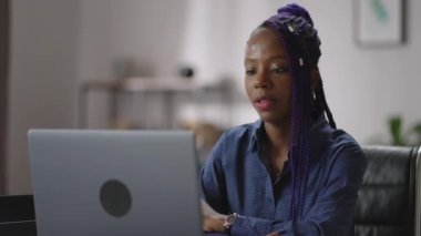 Afro-Amerikalı kadın bilgisayarla iletişim kuruyor, ev ofisinde oturuyor, siyah kadın web kamerasına bakıyor ve ekrana bakıyor.