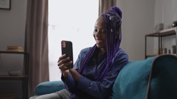 Bella donna nera con acconciatura moderna sta chiamando tramite video chat in smartphone, seduto da solo a casa durante il fine settimana, parlando con gli amici e sorridendo — Video Stock