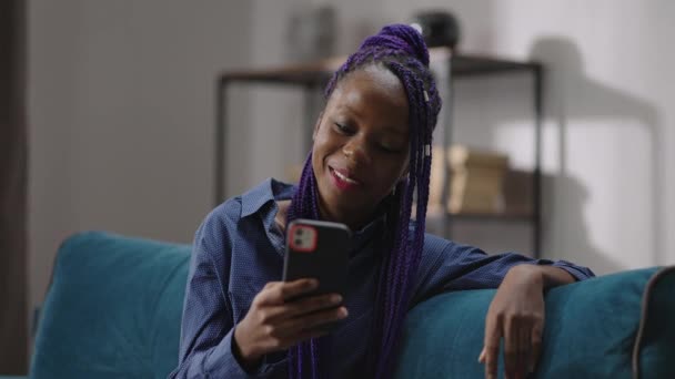 Alegre afro-americana joven mujer está viendo divertido vídeo o película en la pantalla del teléfono inteligente, sentado solo en casa — Vídeos de Stock