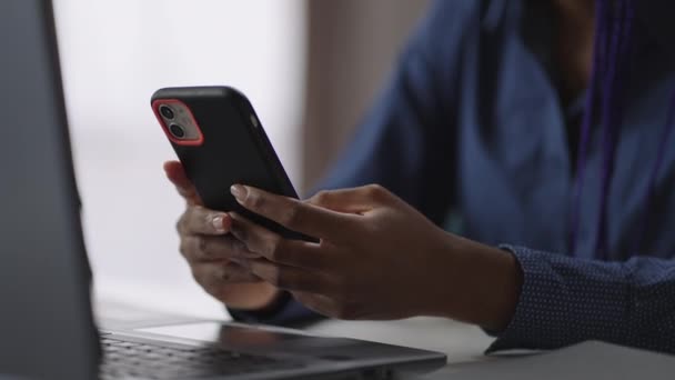 Travailleuse de bureau noire utilise un modèle moderne de smartphone, vérifiant les e-mails et bavardant dans des filets sociaux, assise à table avec ordinateur portable — Video