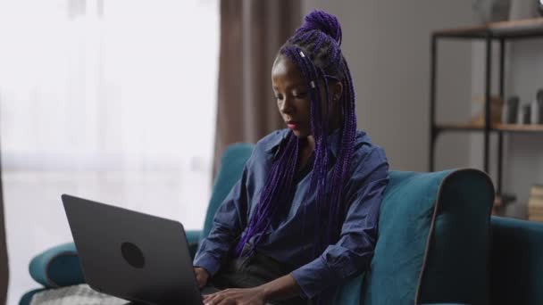 Donna nera adulta sta lavorando con il computer portatile nel fine settimana a casa, seduto sul divano in soggiorno e navigare in internet, chattare nelle reti sociali — Video Stock
