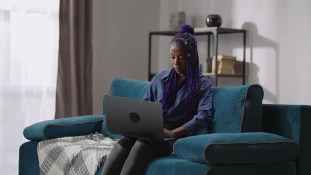 Молодая черная женщина с фиолетовыми дредами работает дистанционно с ноутбуком дома в выходные, внештатная работа для студентов — стоковое видео