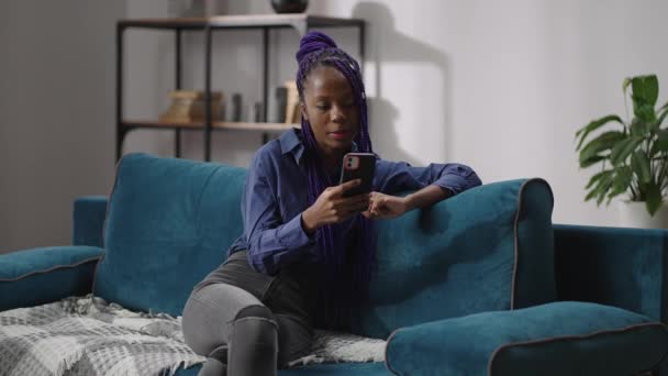 Портрет черной девушки-подростка, общающейся посредством видеочата в смартфоне, дама с дредами сидит на диване в современной квартире — стоковое видео