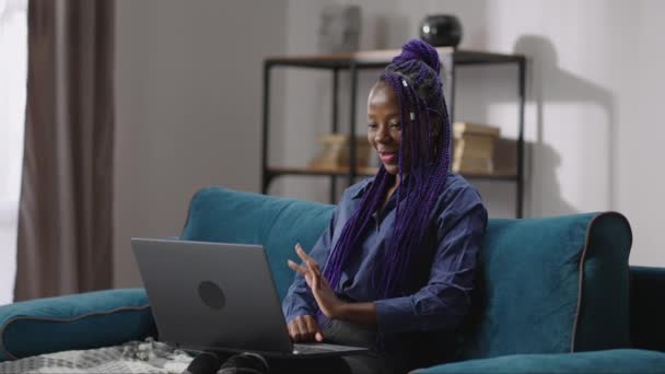 Afro-americano donna sta utilizzando il computer portatile per comunicare tramite fotocamera web, seduto sul divano in camera e sorridente — Video Stock