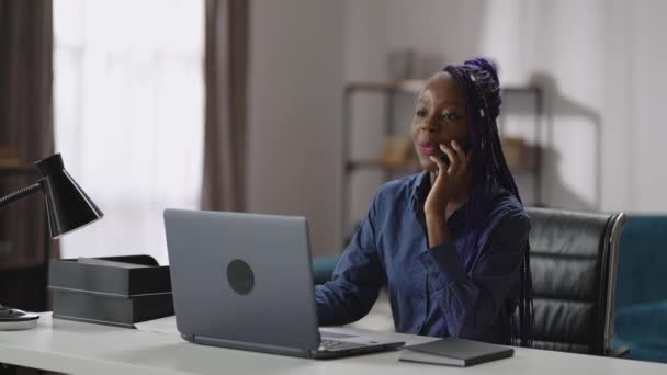 Ung glad svart affärskvinna talar med mobiltelefon i sitt hemmakontor, sitter vid bordet med laptop, chatta med mobiltelefon — Stockvideo