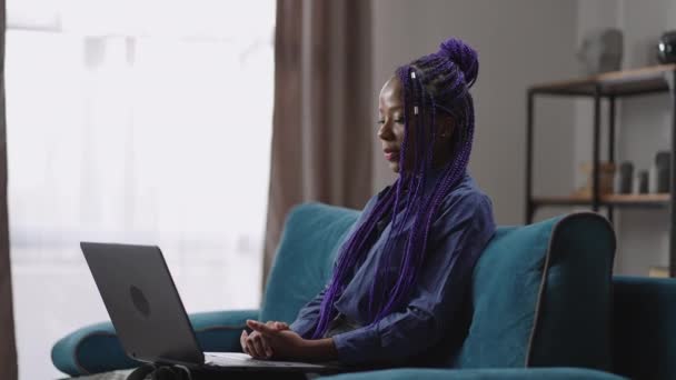 Ung svart kvinna student kommunicerar via online videochatt med bärbar dator, sitter ensam hemma i vardagsrummet — Stockvideo