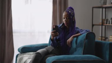 Afro-Amerikalı genç kız kanepesinde tek başına oturuyor, elinde akıllı telefonu tutuyor, dil öğreniyor ya da e-kitap okuyor.