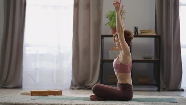 Yogautövande hemma, vuxen kvinna sitter på golvet och sträcker upp händerna, tränar sin kropp — Stockvideo