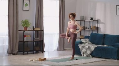 Kadın odada denge için egzersiz yapıyor, kadın bir bacağının üzerinde duruyor, yoga yapıyor.