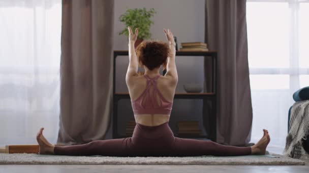 Donna yogi sta facendo split e stretching mani in alto, vista posteriore del corpo femminile in soggiorno, esercizi di yoga e ginnastica — Video Stock