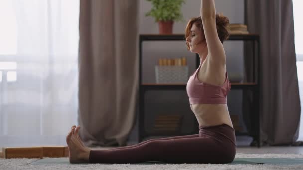 Женщина сидит в позе лотоса на полу в квартире, делает упражнения для растяжения, боковой вид на спортивную женскую фигуру — стоковое видео