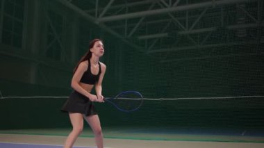 Genç bayan kapalı sahada tenis oynuyor, raketle topa vuruyor, oyunun içinde ağır çekimde.