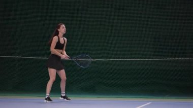 Genç, güzel esmer kadın, tenis oynamayı kapalı sahada öğreniyor. Raketle topa vuruyor. Yavaş çekimde, uzun çekimde.