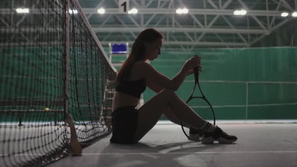 Joven jugadora de tenis está sentada cerca de la red en la cancha después del partido perdido, haciendo girar la raqueta y pensando en fallar — Vídeos de Stock