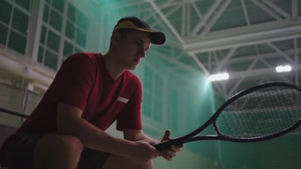 Nervous tennis player is sitting on bench on court before important game, twisting his racquet in hands, medium portarit indoor — Stock Video