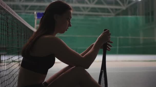 Triste jugadora de tenis está sentada cerca de la red en la cancha, hilando raqueta y pensando en el fracaso, el entrenamiento o la competencia — Vídeos de Stock