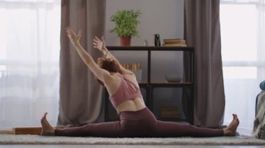 Evde spor yapan esnek bir kadının yoga egzersizi var. Kadın ikiye bölünmüş ve ellerini uzatıyor. Oturma odasında yerde oturuyor.