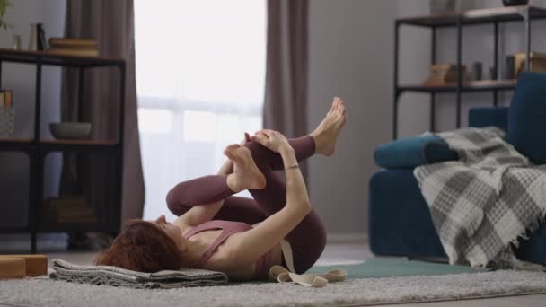 Vrouw doet lichamelijke oefening voor flexibiliteit, met behulp van tape voor het strekken van been, liggend op de vloer in de woonkamer, gezonde levensstijl — Stockvideo
