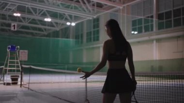 Spor giyinmiş bir kadın tenis kortunun üzerinde yürüyor, elinde tenis raketi ve top atıyor, ince kadın figürünün arka görünüşü.