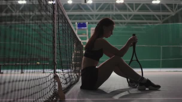 Jugadora de tenis pensativa está sentada en el suelo en la cancha de tenis después de un partido o entrenamiento, torcer la raqueta — Vídeos de Stock