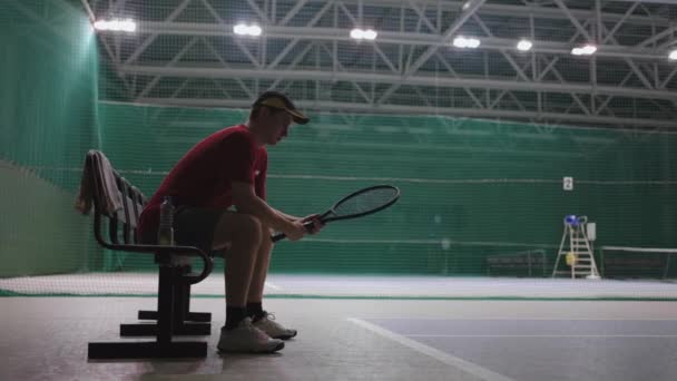 Joueur de tennis masculin est assis sur le banc dans le court de tennis après l'entraînement ou le match, résidant et tordant raquette à la main — Video