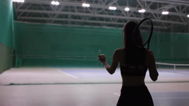 Joven morena está caminando en la cancha de tenis en el interior, lanzando la pelota y la captura, preparándose para servir — Vídeos de Stock