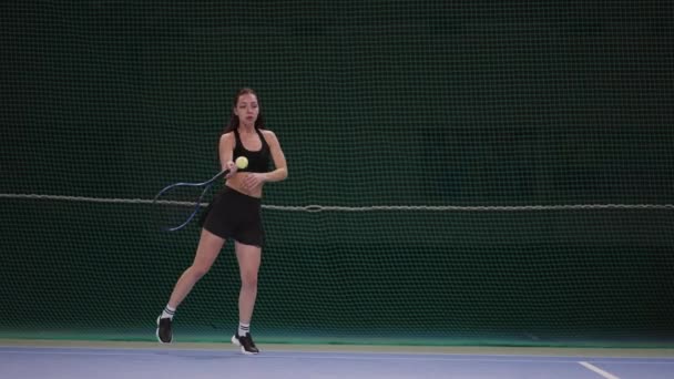 Atraente jogador de tênis feminino delgado está treinando na quadra interna, batendo bola por raquete, tiro de comprimento total em câmera lenta — Vídeo de Stock