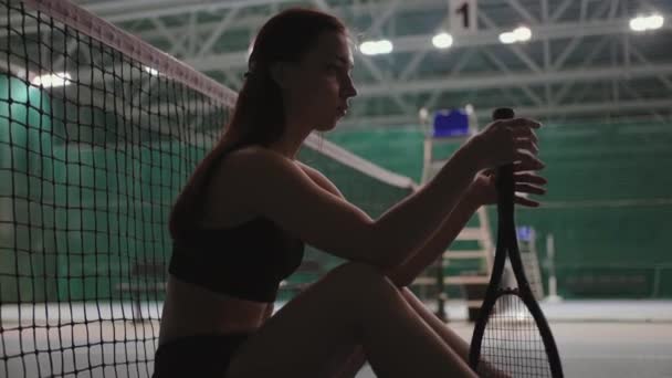 Young pretty female tennis player is sitting on floor on tennis court after match or training, twisting racquet — Stock Video
