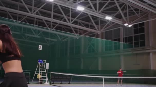 男性と女性が屋内コートで一緒にテニスをしている若いスポーツ女性がテニスラケットでボールを打つ、フル長さのスローモーションショット — ストック動画