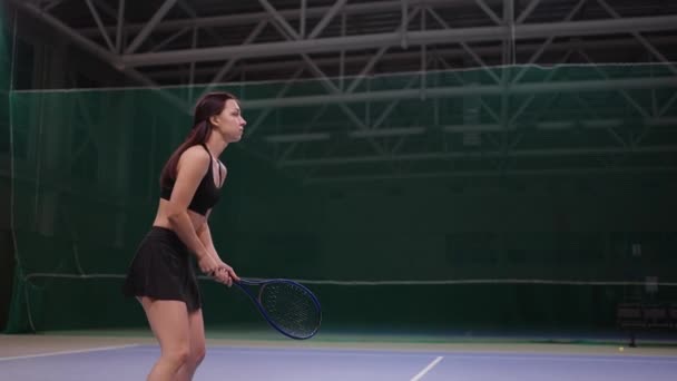 Молодая женщина в спортивной одежде играет в теннис на крытом корте, спортсменка бьет мяч теннисной ракеткой — стоковое видео