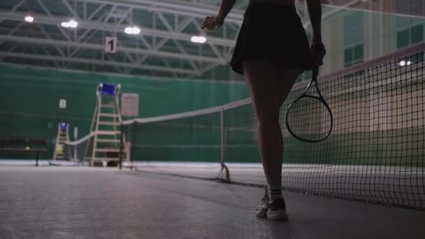Confident sporty woman is walking over tennis court, holding tennis racquet and throwing ball, view on slender trained legs — Stock Video