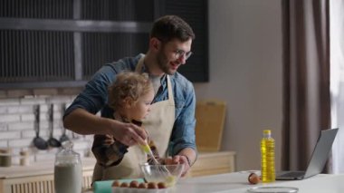 Küçük oğlumla babam dizüstü bilgisayarda ders izliyorlar, mutfakta yemek pişiriyorlar, kasede yumurta çırpıyorlar, baba ve çocuk.