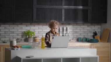 Ciddi bebek aşçı gibi giyinmiş. Mutfakta dizüstü bilgisayarla internette sörf yapıyor.