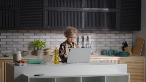 Bebé serio está vestido como un cocinero está de pie en la cocina del hogar, navegar por Internet por el ordenador portátil — Vídeos de Stock