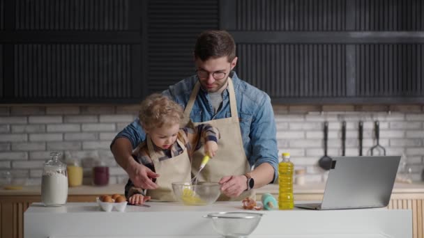 Сімейний час в неділю вранці на кухні, людина вчить свого сина готувати, малюк збиває яйця в мисці, перемішуючи за допомогою пива — стокове відео