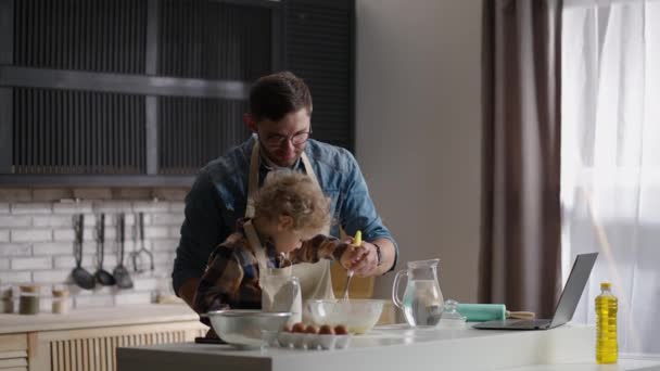 Man en zijn zoon zijn pannenkoeken aan het bakken op zondagochtend, kleine jongen probeert pannenkoekenbeslag te roeren in kom, vader en kind — Stockvideo