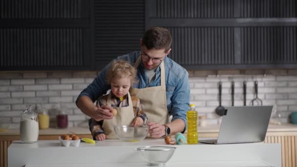 Papa en zoontje brengen tijd samen door in de keuken, het kind studeert om te koken, breekt eieren in een kom — Stockvideo