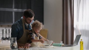 Küçük çocuk babasıyla yemek pişirirken sofra takımıyla oynuyor. Mutlu aile hafta sonları birlikte vakit geçiriyor.