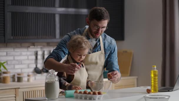 Ευτυχισμένος νεαρός πατέρας και ο μικρός γιος μαγειρεύουν πρωινό μαζί το πρωί του Σαββατοκύριακου, σπάζοντας τα αυγά σε μπολ — Αρχείο Βίντεο