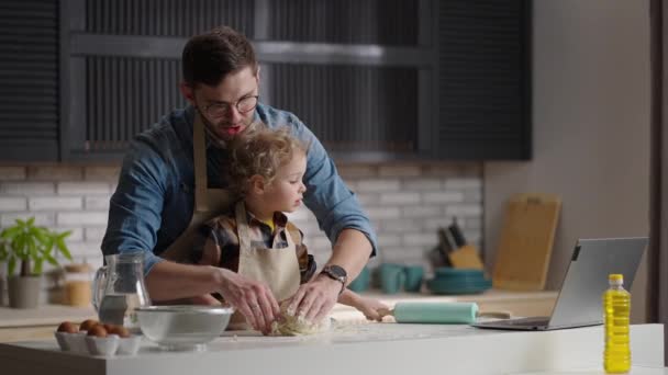 O pai novo com seu filho pequeno está cozinhando, amassando a massa na mesa da cozinha no apartamento moderno — Vídeo de Stock