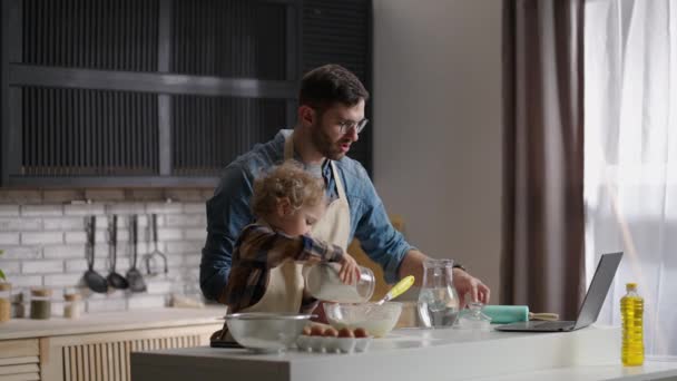 Jovem e seu filho filho estão cozinhando juntos em casa, lendo receita na internet por laptop — Vídeo de Stock