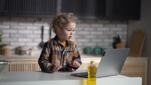 Menino bonito está vendo desenhos animados e jogos na tela do laptop, usando notebook na cozinha em casa no fim de semana — Vídeo de Stock