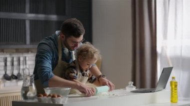Genç baba, küçük oğlunun yemek pişirmesini öğreniyor. Modern apartmandaki mutfak masasında hamuru yuvarlıyor.