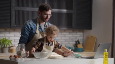 Adam ve oğlu internet tarifine göre yemek pişiriyor, adam laptopta bilgi okuyor ve yeni yürümeye başlayan çocuk hamurla oynuyor.