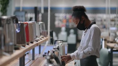 Yüz maskeli genç ev hanımı dükkandaki küçük ev aletlerini seçiyor. Afro-Amerikalı kadın elektrikli su ısıtıcısını izliyor.