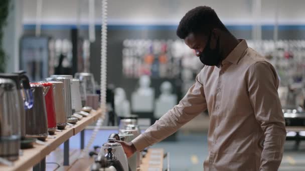 Hombre afroamericano está viendo hervidores eléctricos en el estante de la tienda de electrodomésticos, comprador está usando mascarilla — Vídeos de Stock