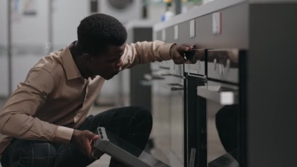 Jeune homme noir regarde four pour la maison, faire du shopping dans le magasin d'appareils ménagers, célibataire choisit l'équipement — Video