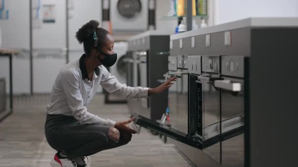 Jeune dame noire choisit cuisinière dans le magasin d'appareils ménagers, portant un masque pour la protection contre le covide-19, en comparant différents modèles — Video