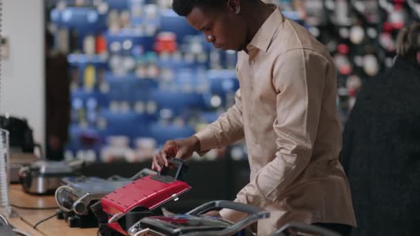 Afro-amerikaanse man is het kiezen van elektrische wafel ijzer in huishoudelijke apparaten winkel, het bekijken van de tentoonstelling monster op de plank — Stockvideo
