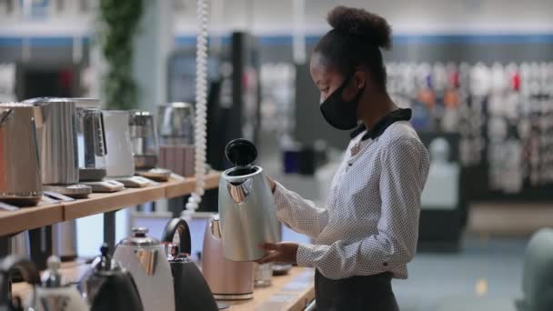 Afro-américaine dame avec masque visage regarde bouilloire électrique dans le magasin d'appareils ménagers dans le centre commercial, le choix de l'appareil pour la cuisine — Video